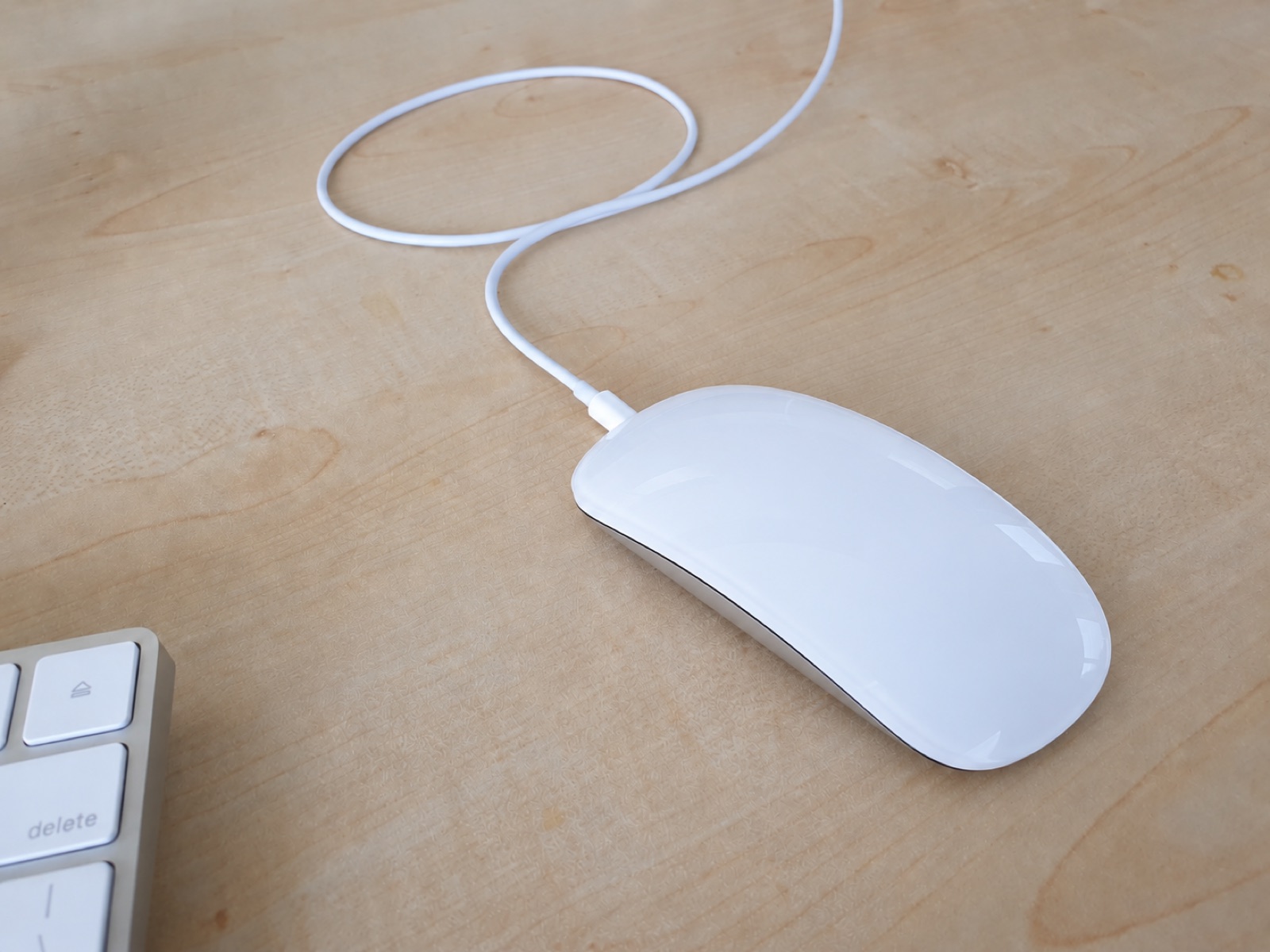 The same wireless mouse sitting upright on a wooden desk next to a keyboard, with the charging cable plugged into the front edge so it can be used while charging.