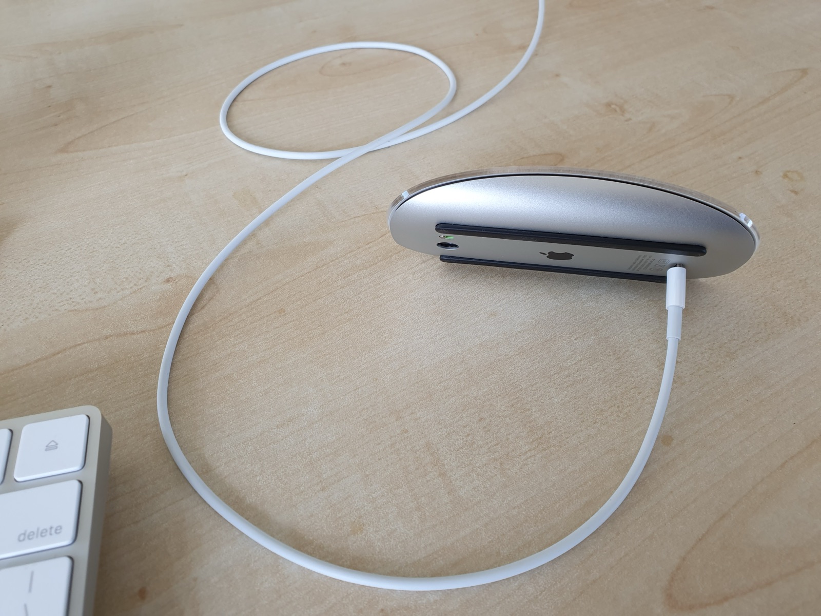 A wireless mouse lying on its side on a wooden desk with a cable plugged into its underside, so it cannot be used while charging.